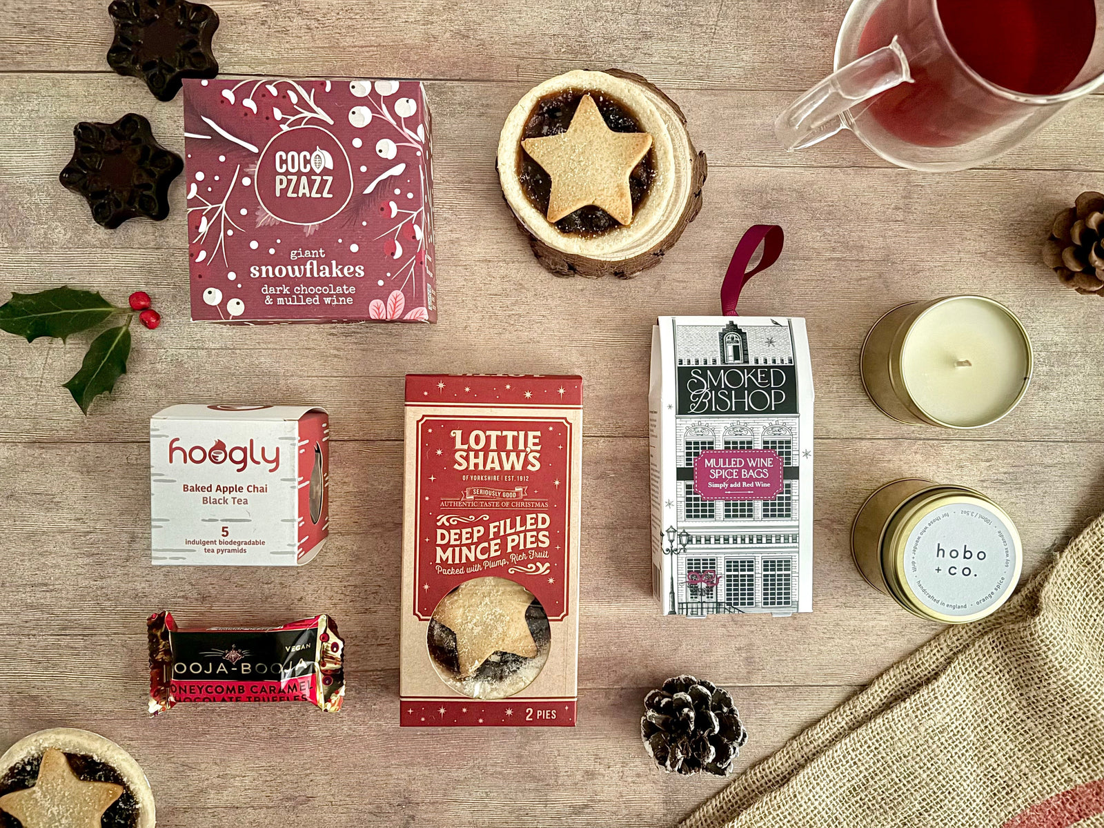 Secret santa festive gifts on flatlay wooden brown background with mince pies, mulled wine dark chocolate snowflakes, mulled wine spice bags, baked apple chai tea pyramids, orange spice candle and honeycomb truffles. Vegan and can be gluten free