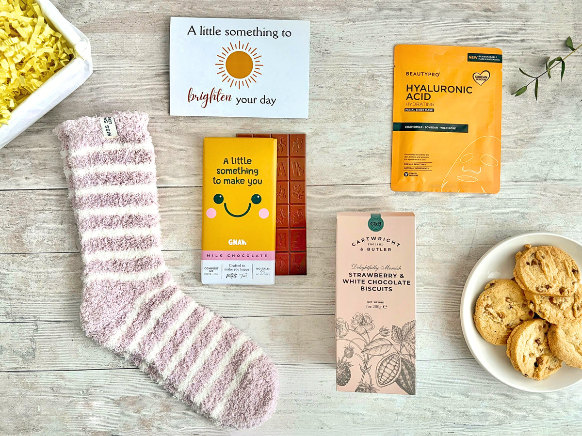 A Little Smile Flatlay on white board with pink and white striped fluffy socks, a chocolate bar with a yellow wrapper with a smile on, a hydrating sheet face mask in yellow packet and pink strawberry & white chocolate biscuits. Presented with a 'A litttle something to brighten your day' card