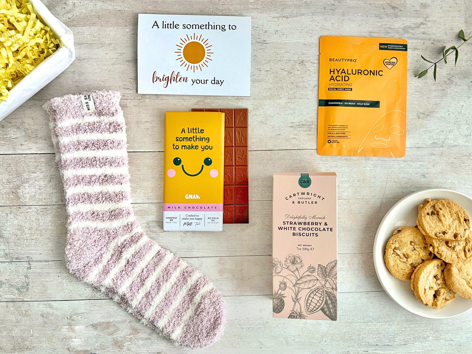 A Little Smile Flatlay on white board with pink and white striped fluffy socks, a chocolate bar with a yellow wrapper with a smile on, a hydrating sheet face mask in yellow packet and pink strawberry & white chocolate biscuits. Presented with a 'A litttle something to brighten your day' card