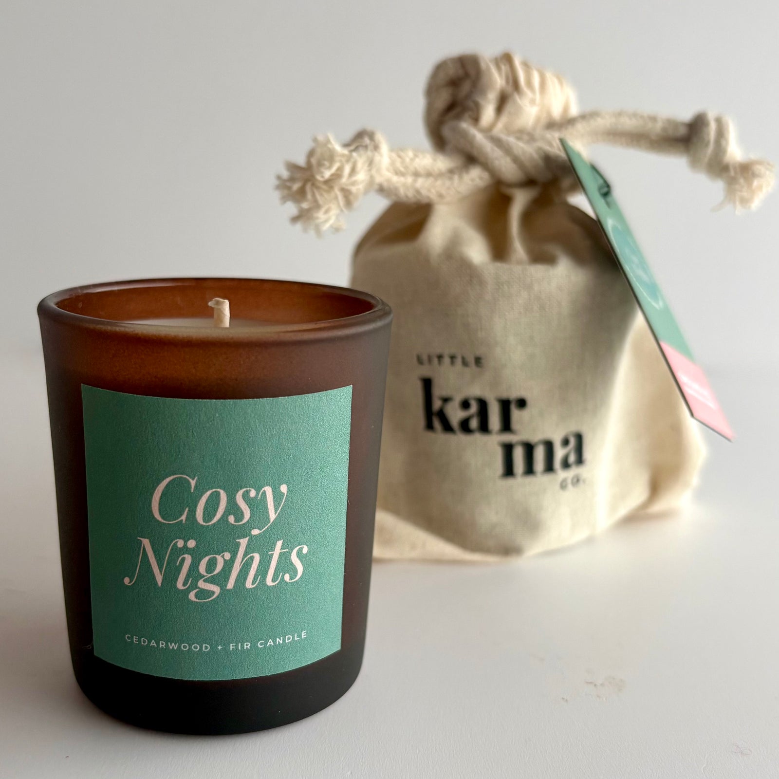 A frosted amber glass candle jar labeled 'Cosy Nights' with a mint green label, placed in front of a beige fabric drawstring pouch