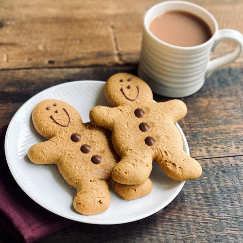 Two gingerbread man cookies decorated with chocolate chips and smiley faces on a white plate, with a cup of coffee on a rustic wooden table in the background.