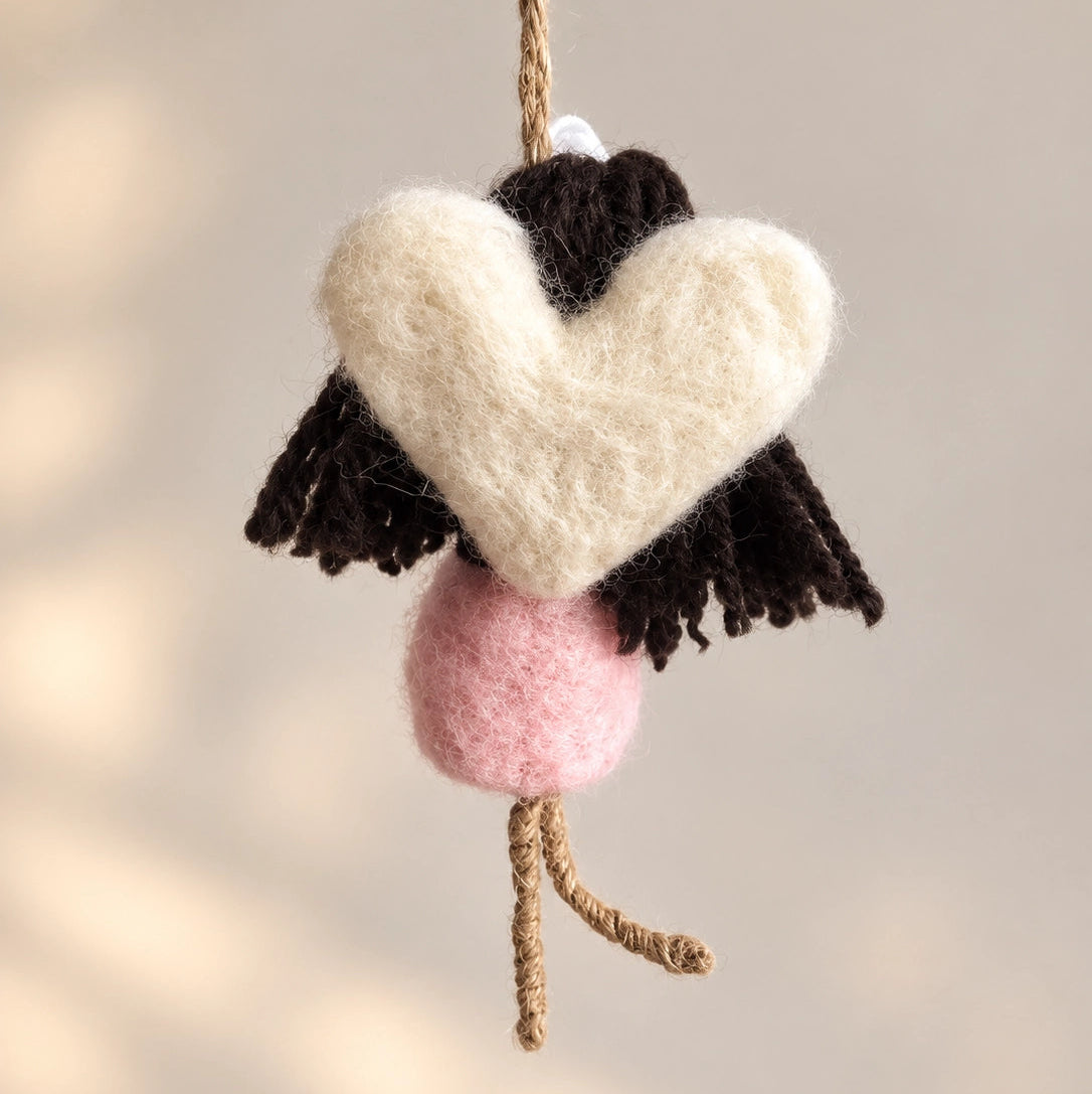 A handmade hanging doll ornament featuring a felted pink dress, dark brown yarn hair, a white fabric flower on its head, and natural jute rope limbs and hanging strap, shown against a soft neutral background.