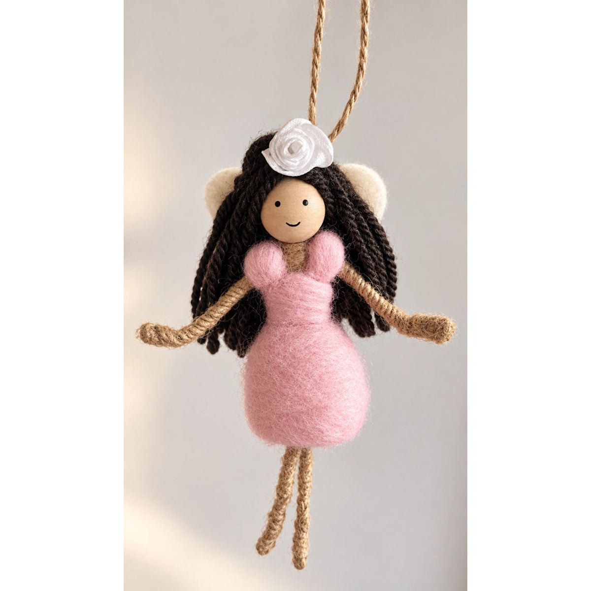 A handmade hanging doll ornament featuring a felted pink dress, dark brown yarn hair, a white fabric flower on its head, and natural jute rope limbs and hanging strap, shown against a soft neutral background.