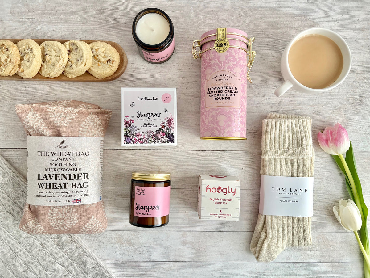 Flat lay with stargazer glass jar candle with pretty floral box, Pink leaf lavender wheatbag, pink tin with strawberry & clotted cream shortbreads, cream alpaca wool bedsocks, Enlglish Breakfast pyramids, Styled with cup of tea and pink and white tulips