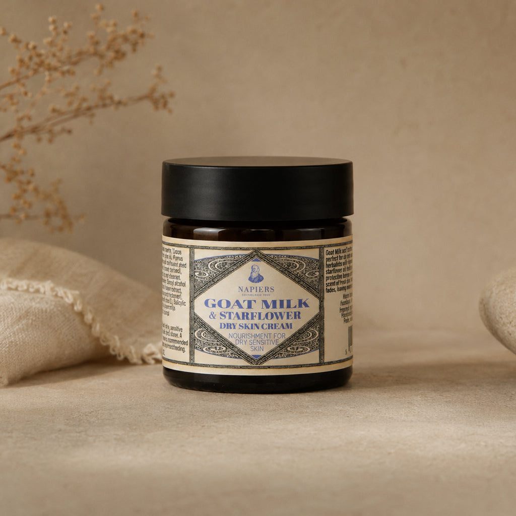 A jar of Napiers Goat Milk & Starflower Ointment stands on a textured beige surface with a linen cloth and dried flowers in the background.