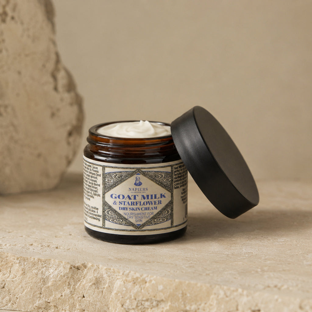 An open amber glass jar of goat milk skin cream with its black lid resting beside it, set on a textured beige stone surface against a neutral beige background.