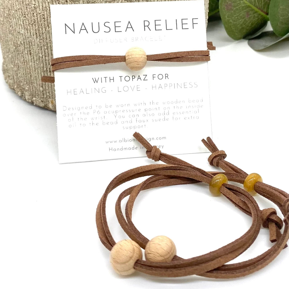 Brown cord anti nausea bracelet with wooden beads and a topaz stone, displayed in front of its branded packaging card on a white surface with a woven basket and green leaves in the background.