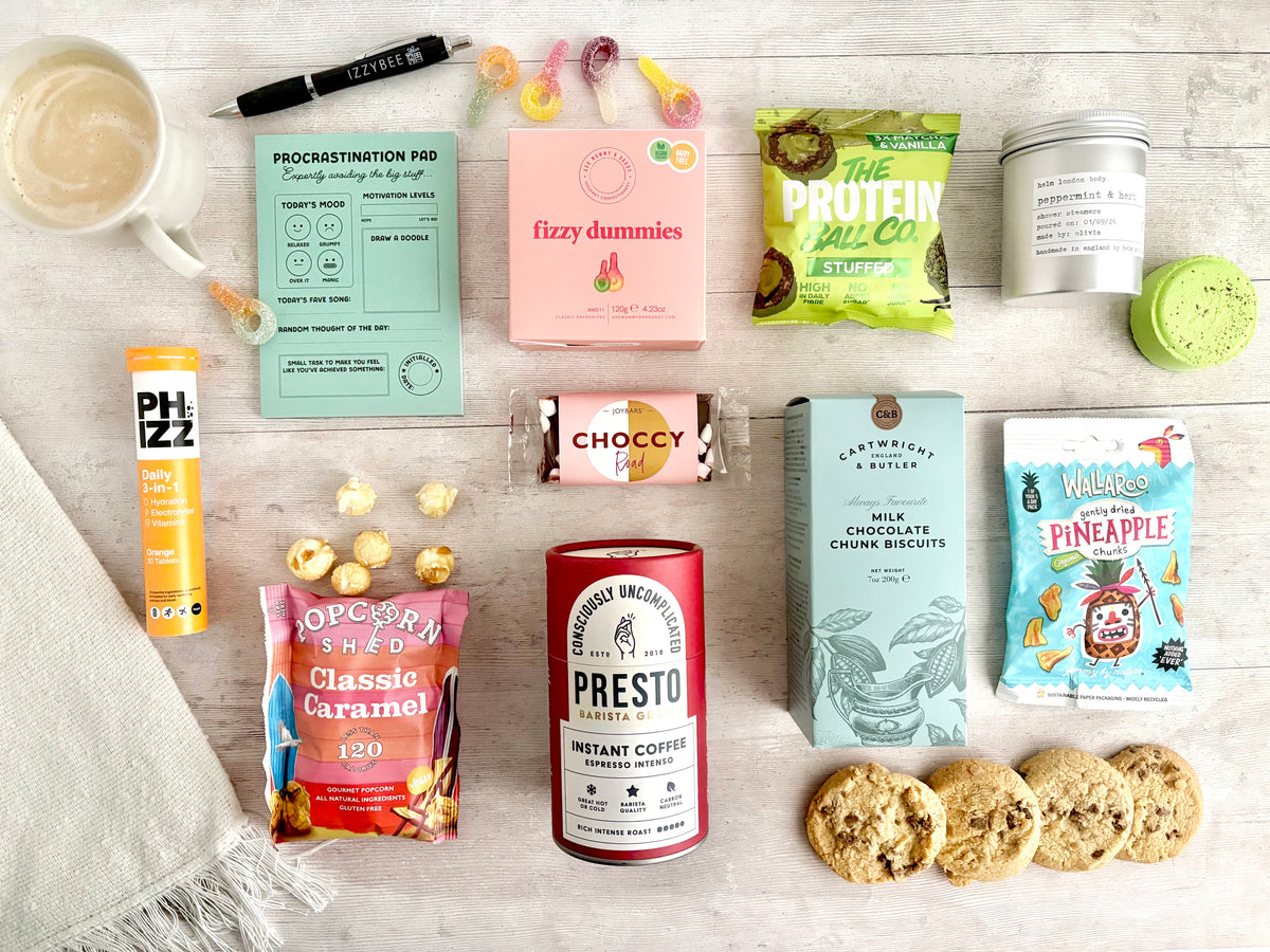 A styled flat-lay of a curated Student Care Package box of snack and coffee gift set on a light wood background, featuring various packaged snacks, a canister of Presto instant coffee, chocolate-covered popcorn, cookies, a pink box labeled 'fizzy dummies', and other small items.