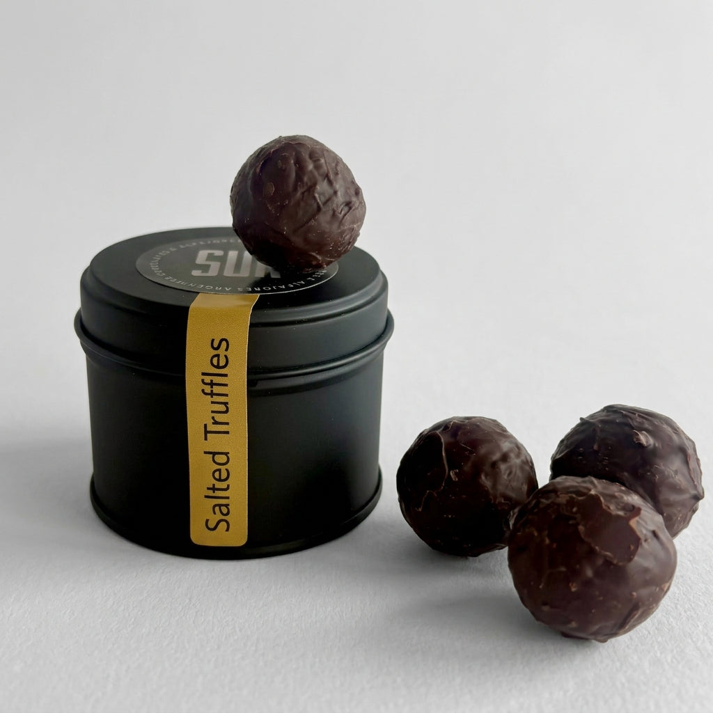A black cylindrical tin of SUR Salted Truffles with a gold label, shown with several chocolate truffles on a light gray surface.