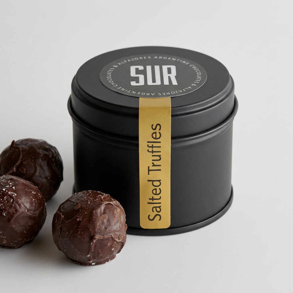 A black cylindrical tin of SUR Salted Truffles with a gold label, shown with several chocolate truffles on a light gray surface.
