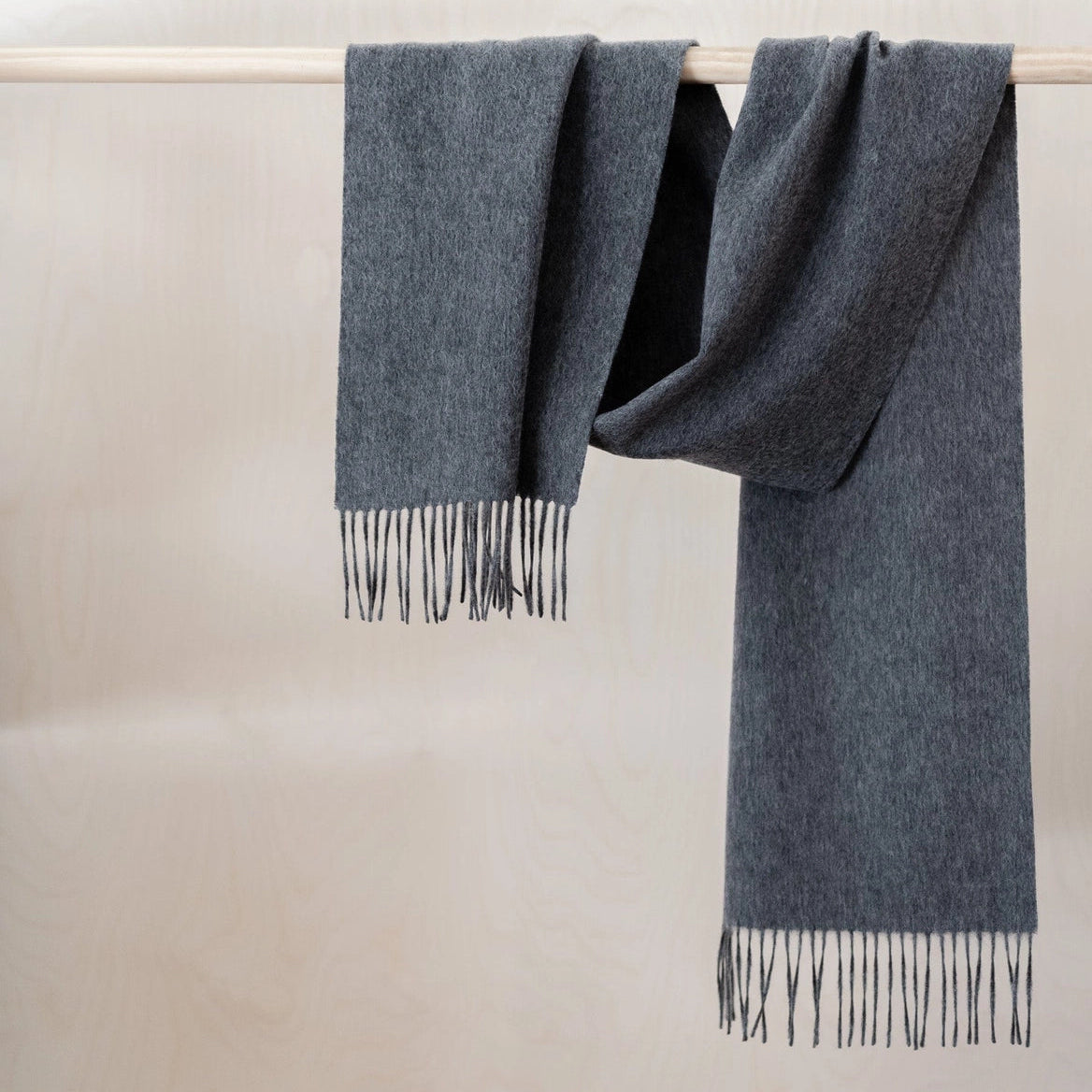 Charcoal Grey 100% lambswool scarf with fringe on wooden rod against a neutral, warm off-white background.