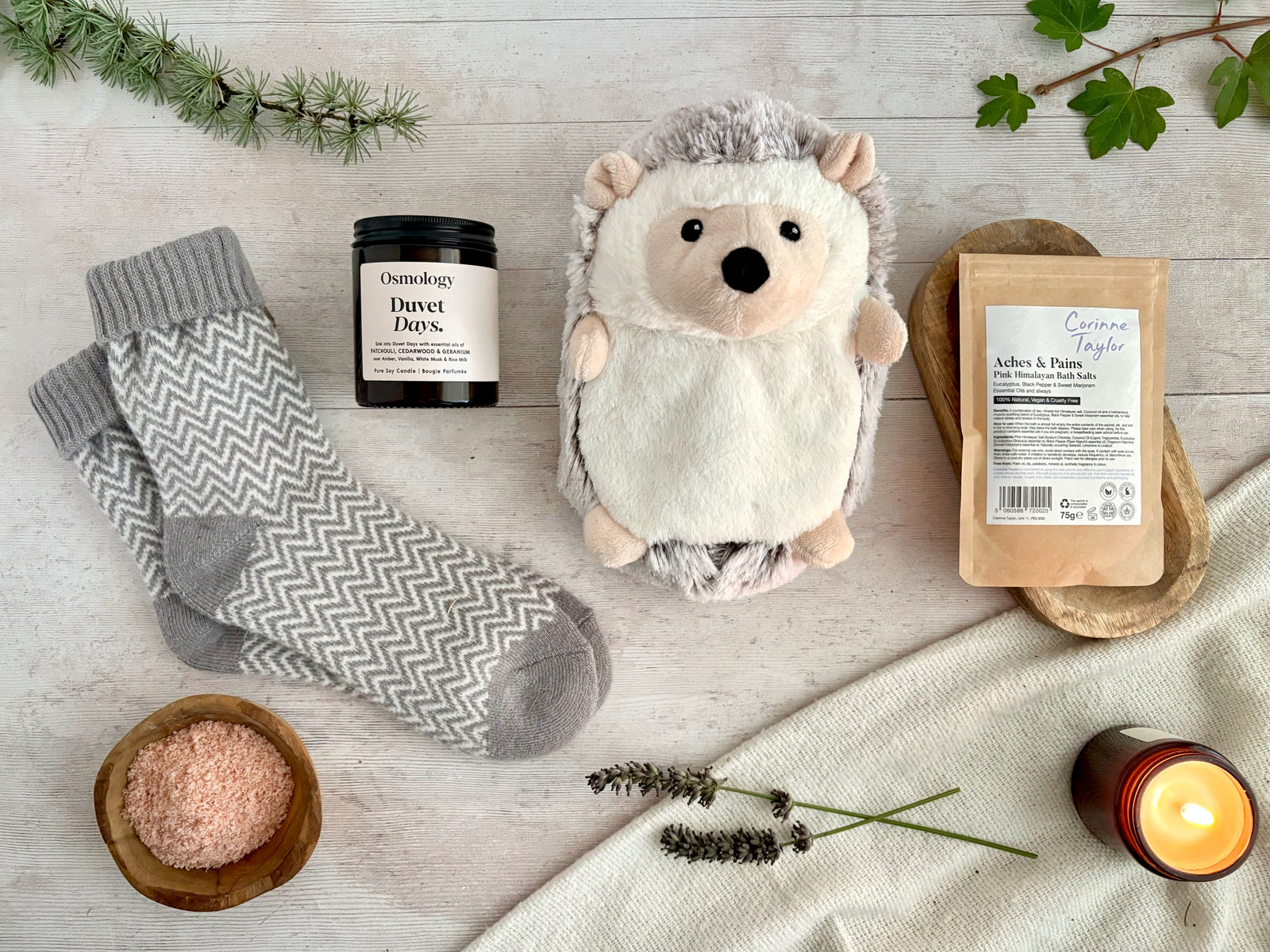 A cosy flat-lay of wellness and home items on a light wood surface: gray patterned socks, a hedgehog plush toy, a candle jar dried lavender sprigs, pine branches, and a lit candle.