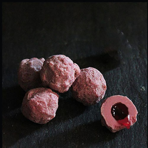 A rectangular pink box of raspberry truffles with a single truffle ball placed in front, featuring the brand 'Willie's Cacao' and a graphic design that includes a fairy and raspberries.