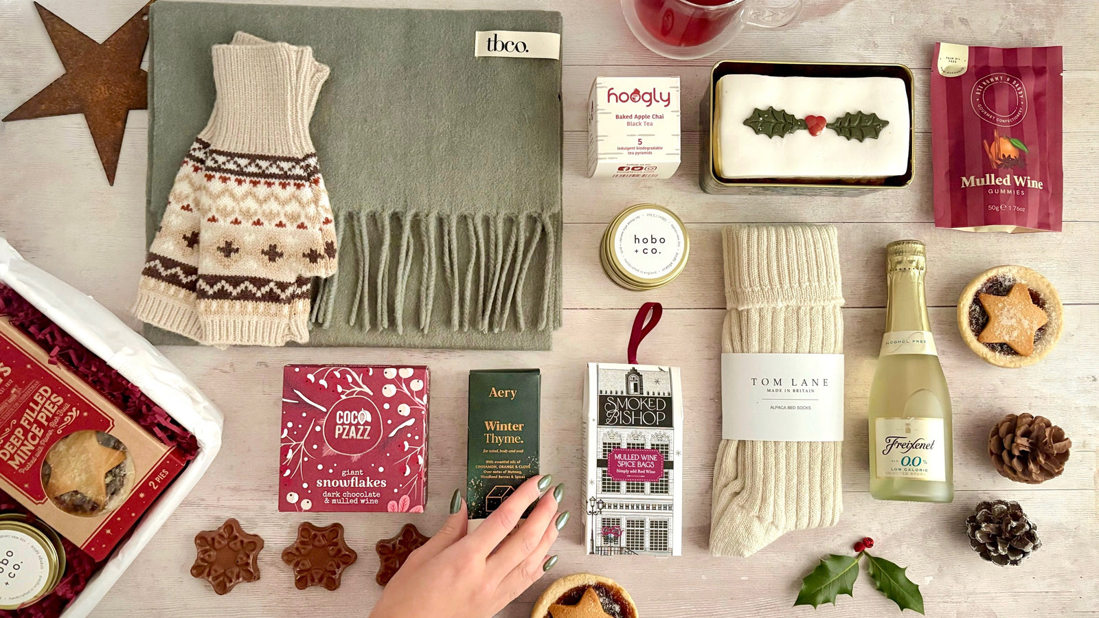 Christmas Gift Hamper Flat Lay with Gift box cosy socks, chocolate snowflakes, candle, christmas cake, mince pies, holly, pinecones, tea and mulled wine spices, scarf and gloves on white wood board