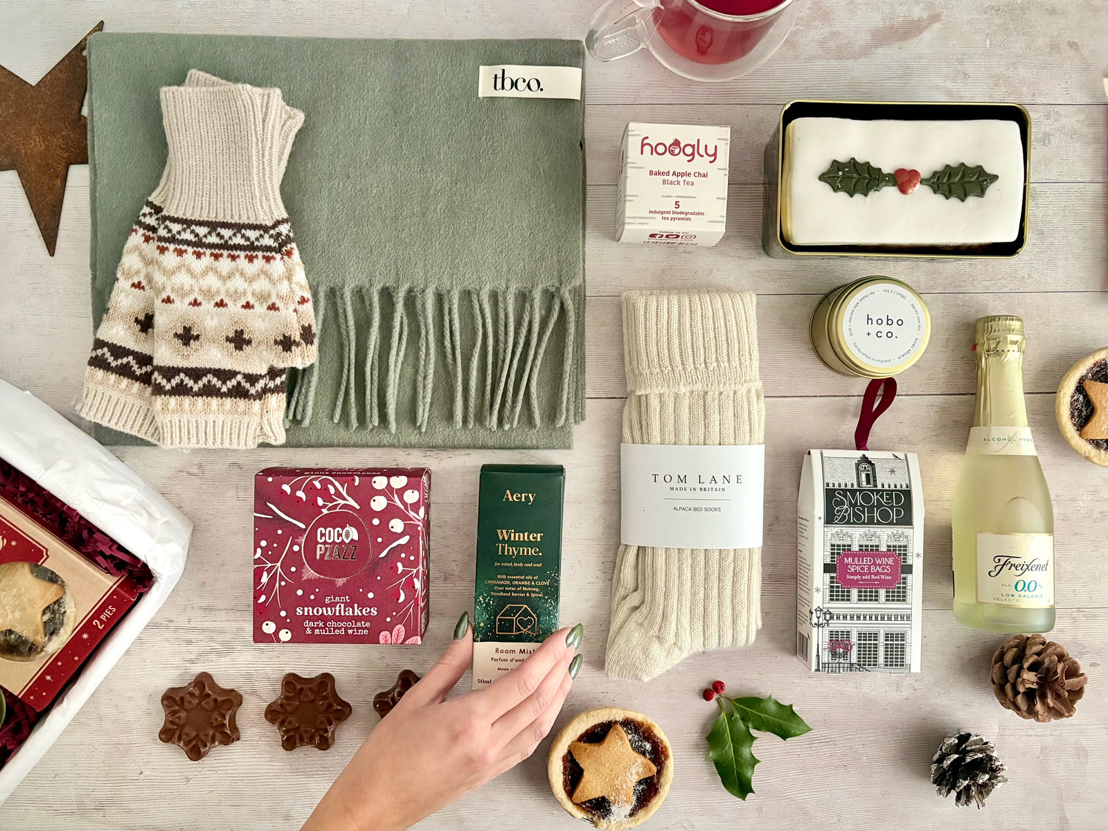 Christmas Gift Hamper Flat Lay with Gift box cosy socks, chocolate snowflakes, candle, christmas cake, mince pies, holly, pinecones, tea and mulled wine spices, scarf and gloves on white wood board